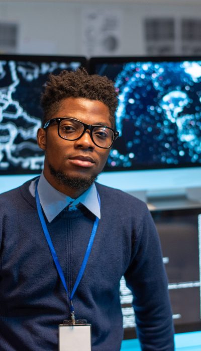 Contemporary young programmer standing by workplace against group of computer monitors with coded data on screens